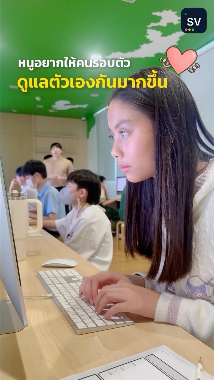 A student in a white sweater types on an iMac in a computer classroom, Thai caption text above her