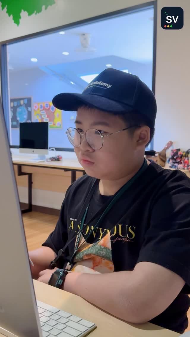 A student in a navy cap and glasses works at an iMac in a bright computer classroom