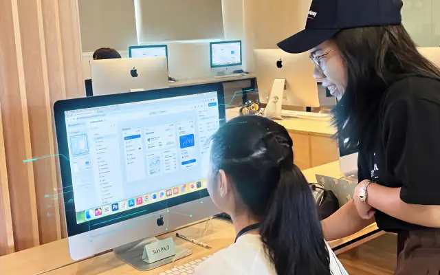 A mentor guiding a student through her app on an iMac