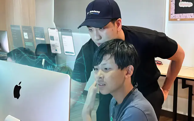 A mentor leaning over to help a student with his app