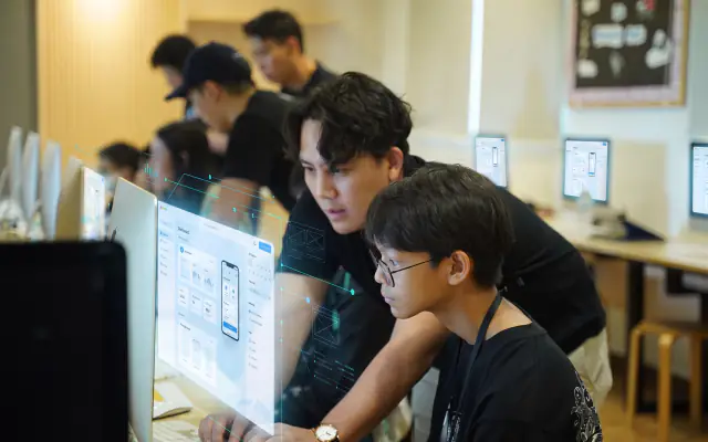 A mentor leaning in to help two students build their app at an iMac