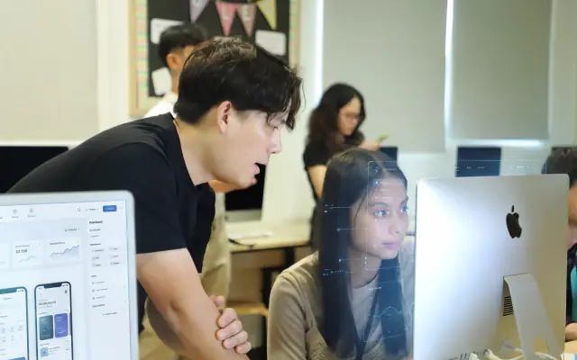A mentor guiding a student as she builds her app on an iMac